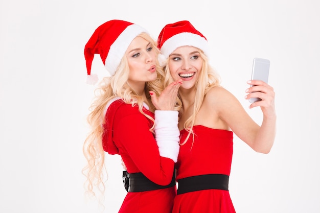 Deux jolies jeunes sœurs jumelles en robes et chapeaux de père Noël rouges montrant un ordinateur portable avec un écran blanc près de l'arbre de Noël sur fond blanc