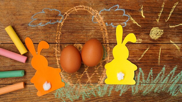 Deux lapins de Pâques en papier et oeufs dans un panier. Dessiné avec de l'herbe de craie colorée et du soleil sur une surface en bois. Dessin festif avec des crayons de couleur.