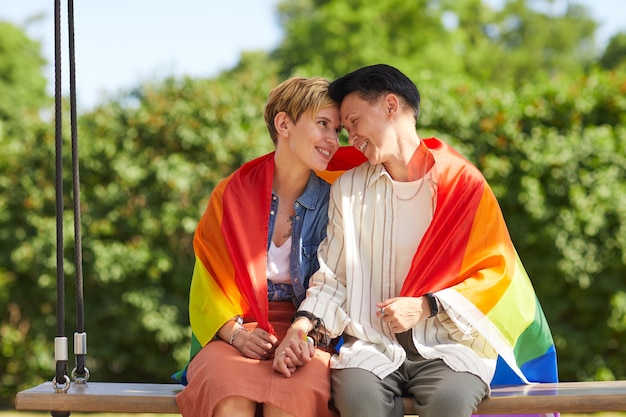 Deux lesbiennes assises sur la balançoire couvrant avec un drapeau lesbien et embrassant à l'extérieur