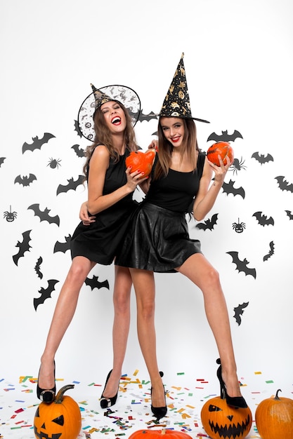 Deux magnifiques filles en robes noires, chapeaux de sorcière et talons hauts s'amusent avec des citrouilles d'halloween.