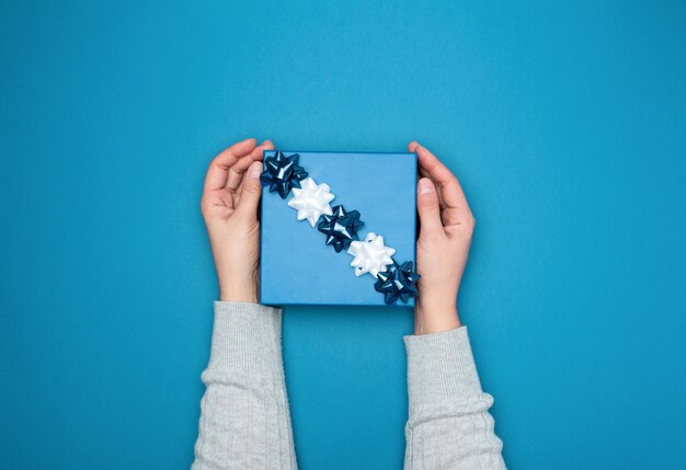 Deux mains féminines dans un pull tiennent une boîte bleue carrée avec un arc sur un fond bleu, vue de dessus, toile de fond festive