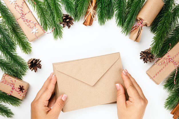 Deux mains de femme tiennent une enveloppe en papier, des décorations de branches d'arbres de Noël et des coffrets cadeaux en arrière-plan.