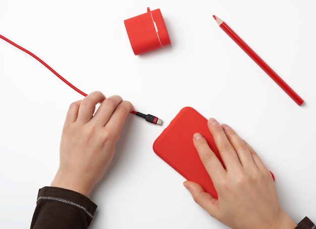 Deux mains tenant un smartphone mobile rouge et un câble de chargement sur une surface blanche