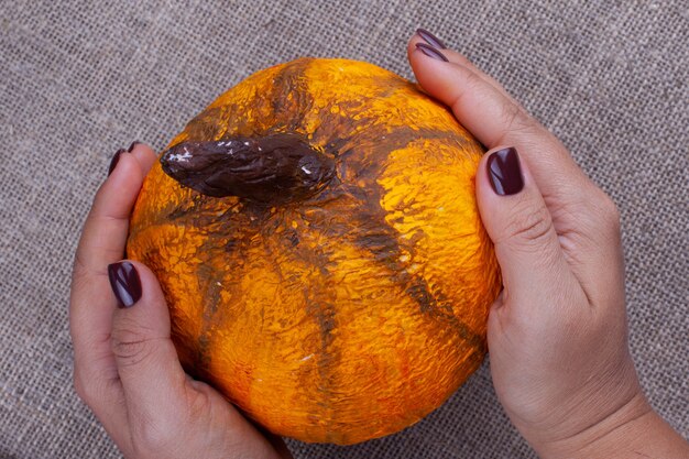 Deux mains tiennent une citrouille en papier mâché orange maison pour Halloween sur une surface de jute, vue du dessus