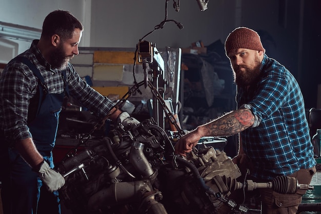 Deux mécaniciens barbus réparent le moteur de la voiture qui est soulevé sur l'élévateur hydraulique du garage. Station-service.