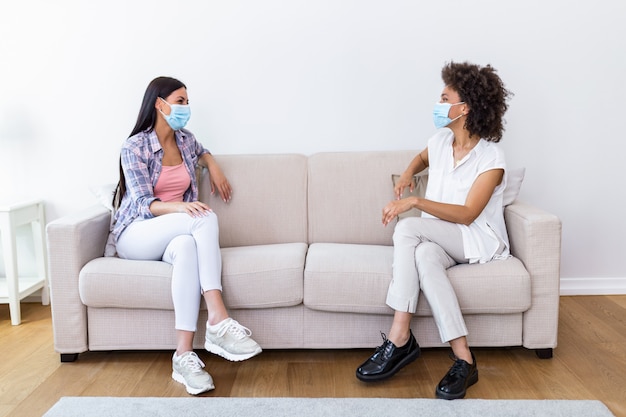 Deux meilleures amies assises à distance sociale portant un masque facial et parlant sur le canapé, empêchant la propagation de l'infection par le coronavirus Covid 19.