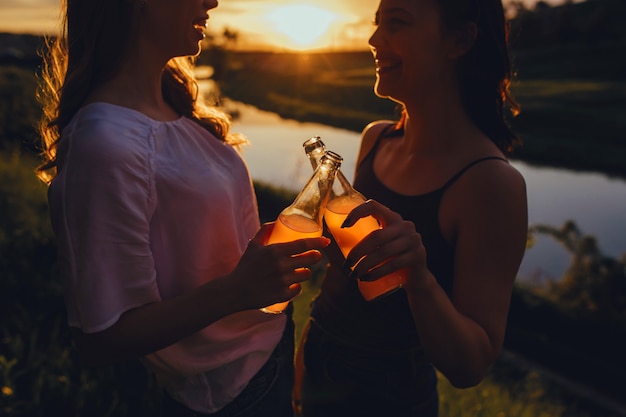 Deux meilleurs amis tenant une bouteille, les filles s'amusant avec un cocktail au coucher du soleil, l'expression du visage positive, en plein air, le concept de la joie