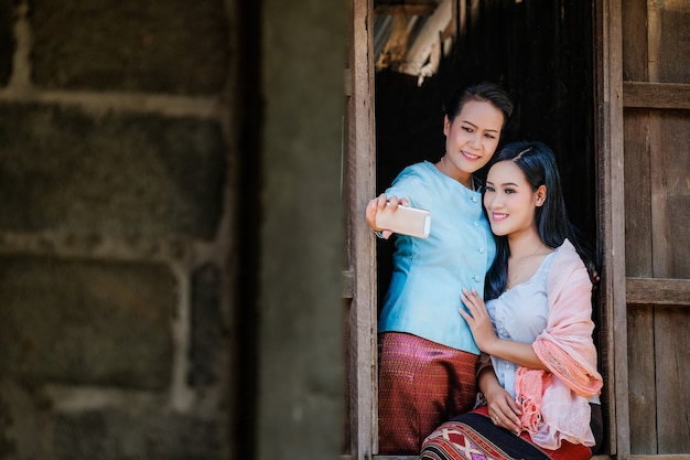 Deux mère et fille vêtues d'une robe thaïlandaise traditionnelle prennent des photos d'elles-mêmes avec un téléphone portable depuis une fenêtre en bois.