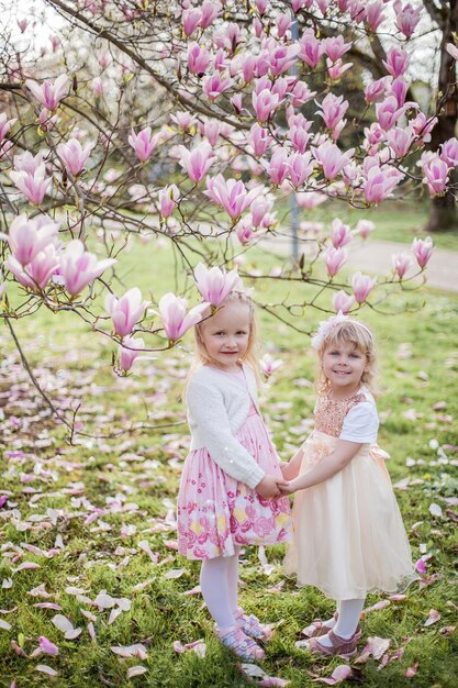 Deux mignonnes petites filles blondes de 3 ans jouent dans le parc près d'un magnolia en fleurs. Boire du thé. Pâques. Printemps.