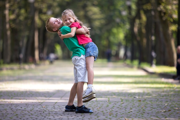 Deux mignons jeunes enfants souriants drôles, fille et garçon, frère tenant sœur dans ses bras, s'amusant sur les arbres verts floues et ensoleillées du parc bokeh. Concept de relations de frères et sœurs aimants.