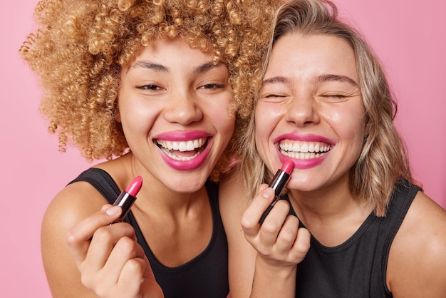 Deux modèles féminins bouclés positifs mettent du rouge à lèvres se préparent pour un rendez-vous ou une fête sourient à pleines dents épaule contre épaule ont des expressions heureuses posent à l'intérieur Cosmétiques pour femmes et procédures de beauté