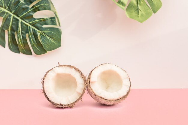 Deux moitiés de noix de coco crue fraîche sur fond rose avec des feuilles de monstera vertes sur blanc. Concept tendance