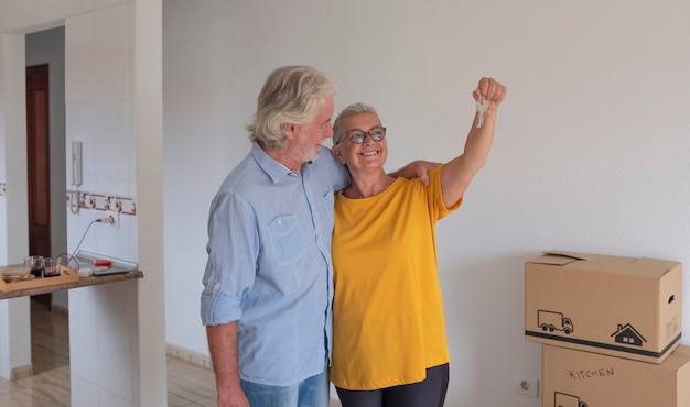 Deux personnes âgées s'embrassant tenant les clés du nouvel appartement vide avec des cartons de déménagement sur le sol - concept de personnes âgées actives et nouveau départ comme à la retraite