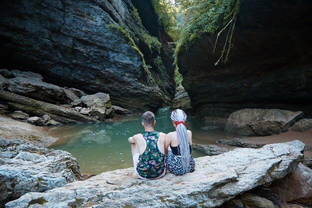 Deux personnes s'assoient sur un gros rocher et regardent la grotte, les rochers et la rivière de montagne, le lac entre les rochers, la forêt tropicale