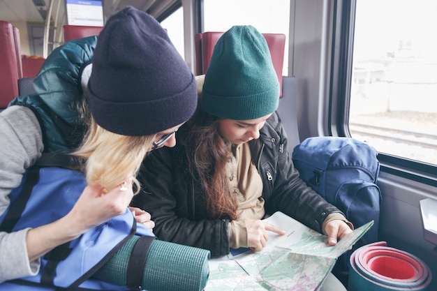 Deux petites amies voyageant avec des sacs à dos et un inventaire vont dans le train et regardent l’itinéraire plus loin sur la carte