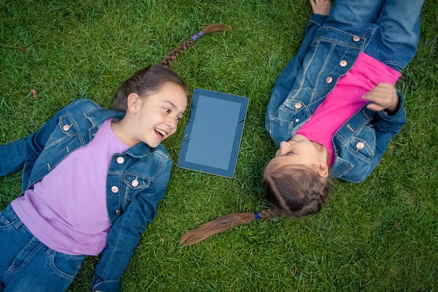 Deux petites filles allongées face à face sur l'herbe et regardant une tablette