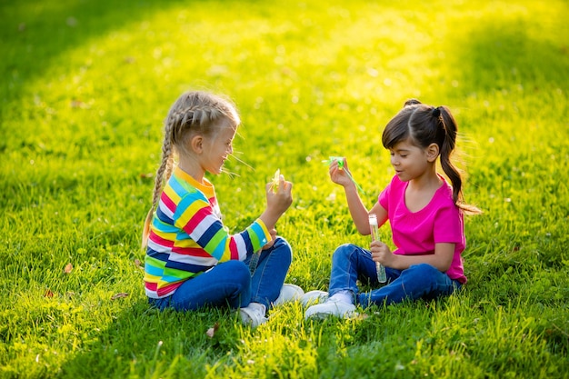 Deux petites filles blondes et brune d'été s'asseoir sur la pelouse soufflant des bulles de savon enfants de l'ethnie européenne et indienne