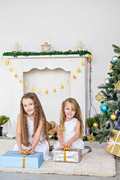 Deux petites filles blondes en robes blanches jouent devant la cheminée et le sapin de Noël avec des boîtes.