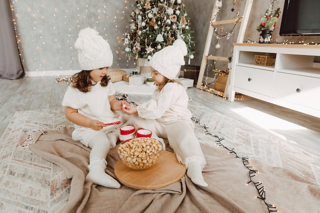 Deux petites filles en chapeaux tricotés d'hiver sont assises sur le sol près de l'arbre de Noël et mangent du pop-corn. notion de noël.