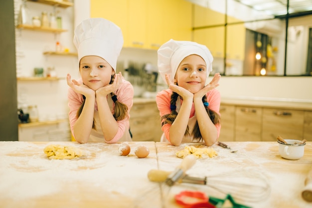 Deux petites filles cuisinières en casquettes sont assises à table, préparation de biscuits dans la cuisine. Enfants cuisinant des pâtisseries, enfants chefs préparant un gâteau