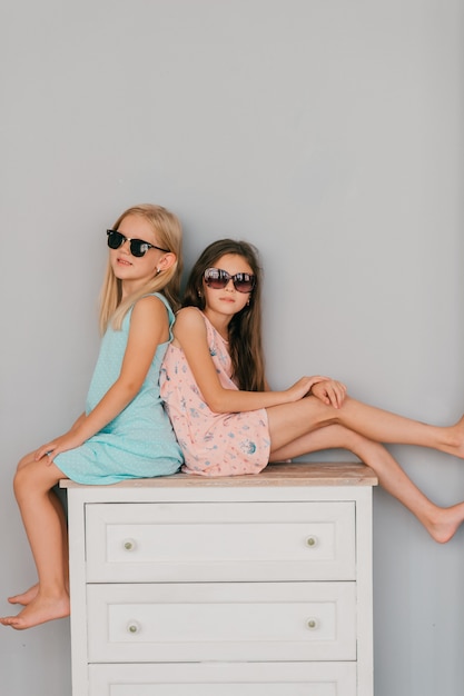 Deux petites filles élégantes en robes colorées et lunettes de soleil avec des visages émotionnels implantation sur commode sur mur gris