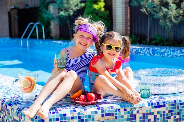 Deux petites filles s'assoient au bord de la piscine en été portant des lunettes de soleil et buvant de la limonade