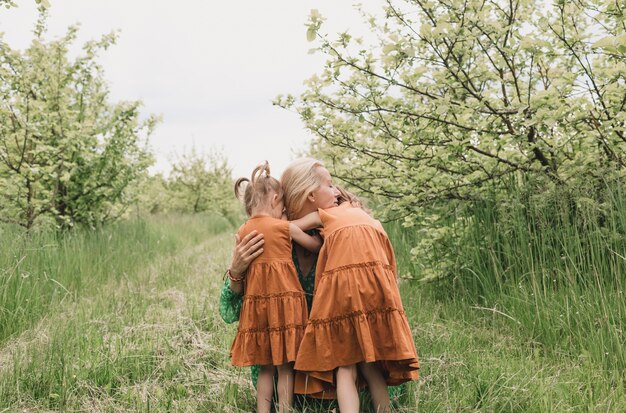 Deux petites filles vêtues de robes identiques embrassent une mère heureuse dans un verger de pommiers