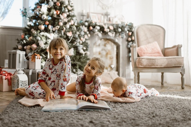 Deux petites soeurs et un petit frère s'allongent sur le tapis et lisent un livre près de l'arbre du Nouvel An