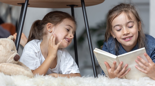 Deux petites sœurs s'amusent à lire un livre ensemble allongées par terre dans leur chambre.