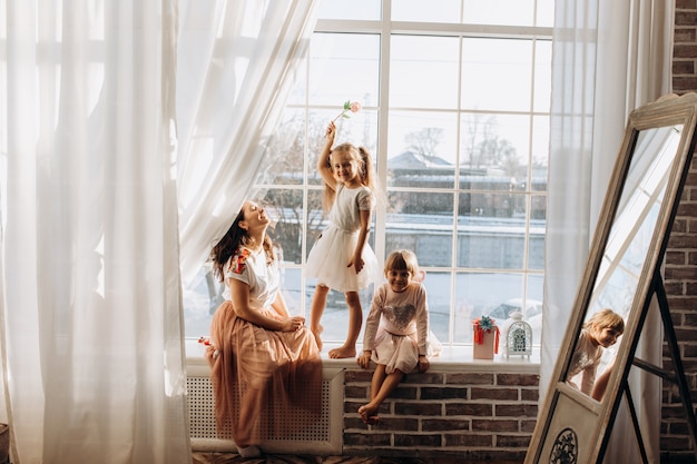 Deux petites sœurs vêtues de belles robes et la jeune mère sont assises sur le rebord de la fenêtre à côté du miroir l'hiver est dehors.