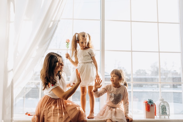 Deux petites sœurs vêtues de belles robes et la jeune mère sont assises sur le rebord de la fenêtre à côté du miroir l'hiver est dehors.