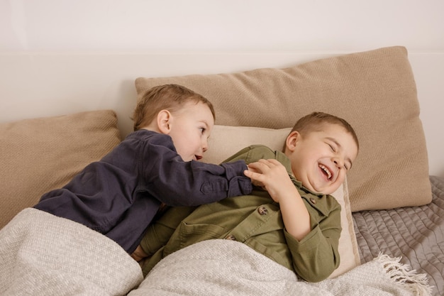 Deux petits garçons caucasiens mignons jouant ensemble sur le lit à la maison. Intérieur et vêtements aux couleurs de la terre naturelle. Environnement cosy. Les enfants s'amusent, deux frères se chatouillent.