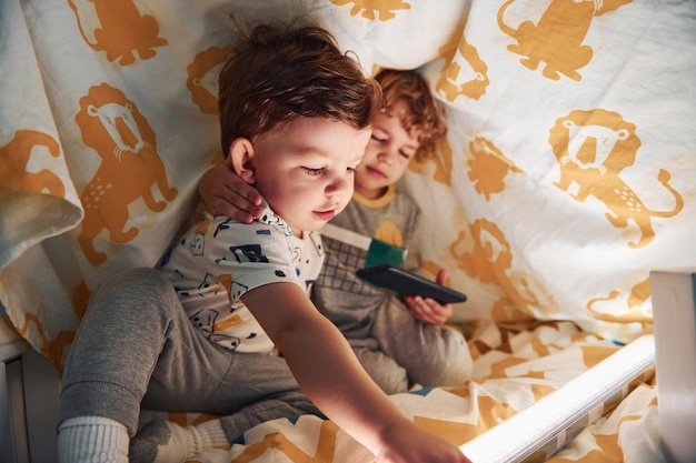 Deux petits garçons se reposent et s'amusent à l'intérieur sous une couverture avec un smartphone