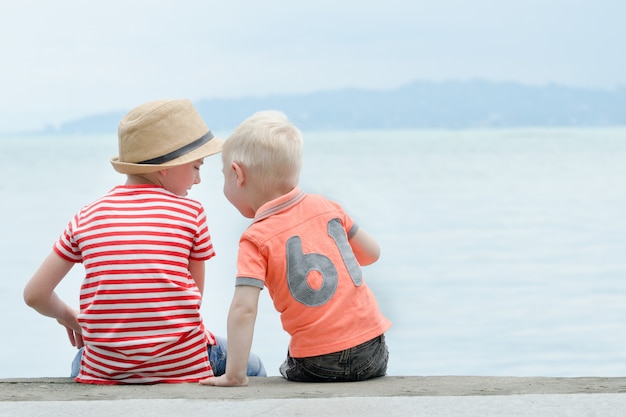 Deux petits garçons sont assis sur une jetée, contre la mer et les montagnes. Vue arrière