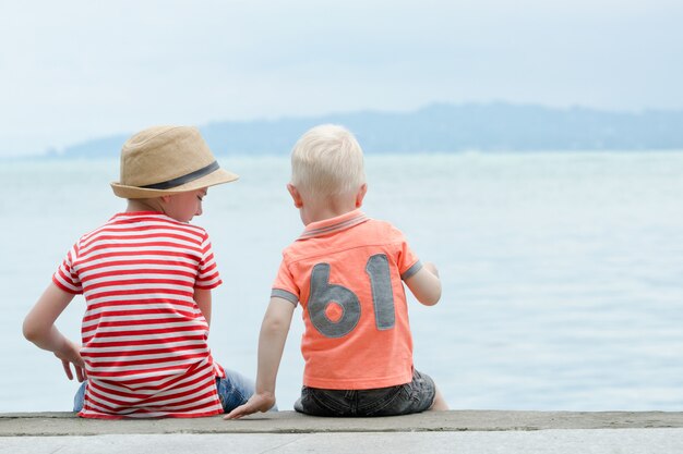 Deux petits garçons sont assis sur une jetée, contre la mer et les montagnes. Vue arrière