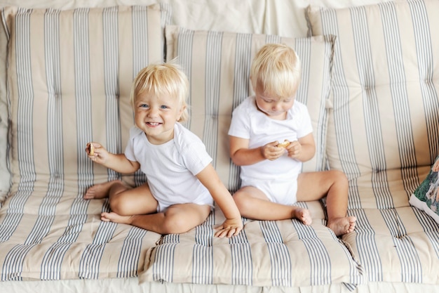 Deux petits jumeaux. Deux bébés identiques vêtus de vêtements blancs pour enfants sont assis sur un canapé dans la chaleur de la maison et tiennent un biscuit à la main. Portrait spontané de jumeaux aux yeux bleus et aux cheveux blonds