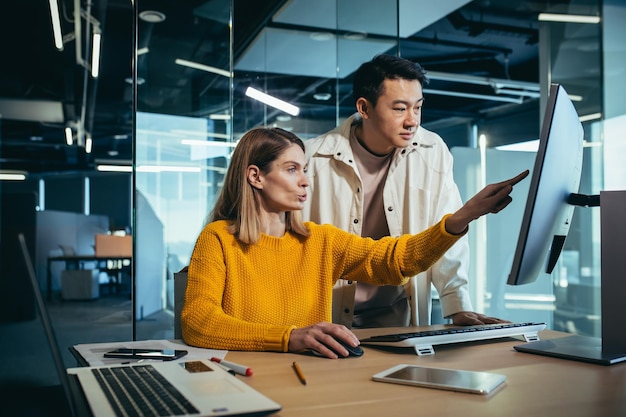 Deux pigistes, un homme asiatique et une femme, travaillent dans un bureau moderne et discutent d'un projet commun en regardant un écran d'ordinateur