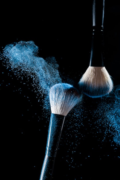 Deux pinceaux de maquillage avec des ombres de maquillage bleu en mouvement sur un fond noir.