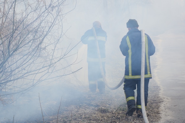 Deux pompiers en fumée bataille incendie de forêt. Un pompier courageux s'enflamme pour éteindre un incendie, un autre pompier tient et porte un tuyau