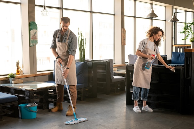 Deux propriétaires de restaurant ou café contemporain lavant le sol et désinfectant les meubles avec un désinfectant après la journée de travail