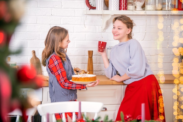 Deux soeurs heureuses décorent la maison de table de Noël dans la cuisine, tenant des tasses et des tartes sucrées dans leurs mains.