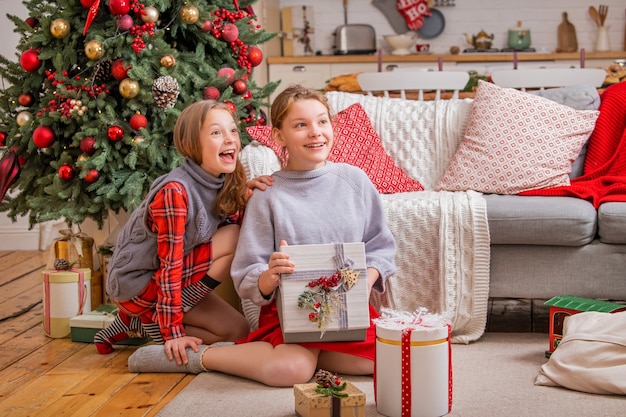 Deux soeurs joyeuses sont assises à la maison près de l'arbre de Noël et regardent des boîtes avec des cadeaux