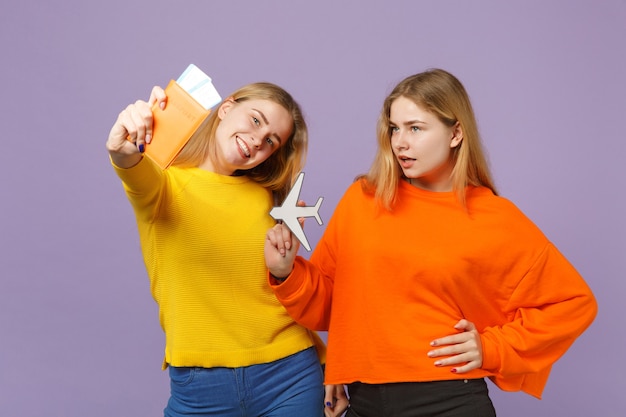 Deux sœurs jumelles blondes souriantes vêtues de vêtements colorés détiennent un passeport, un billet d'embarquement, un avion en papier isolé sur un mur bleu violet. Concept de mode de vie familial de personnes.