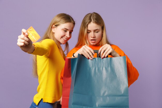 Deux sœurs jumelles blondes souriantes vêtues de vêtements vifs détiennent une carte bancaire de crédit, un sac d'emballage avec des achats après le shopping isolés sur un mur bleu violet. Concept de famille de personnes.