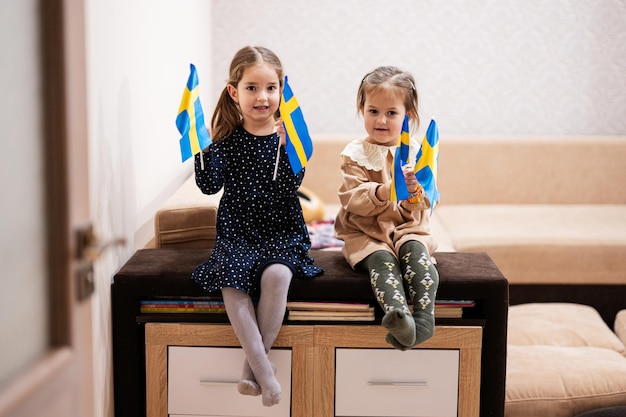 Deux sœurs sont assises sur un canapé à la maison avec des drapeaux suédois sur les mains Suède enfants filles avec drapeau