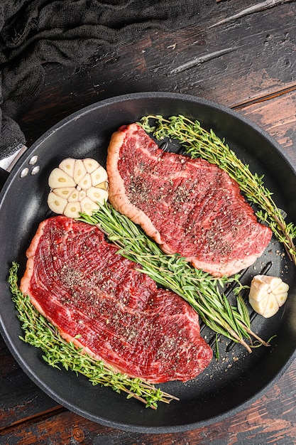 Deux steaks de bœuf bio picanha crus prêts à être grillés sur une poêle avec des herbes et de l'ail. Sur le vieux fond en bois sombre, vue du dessus.