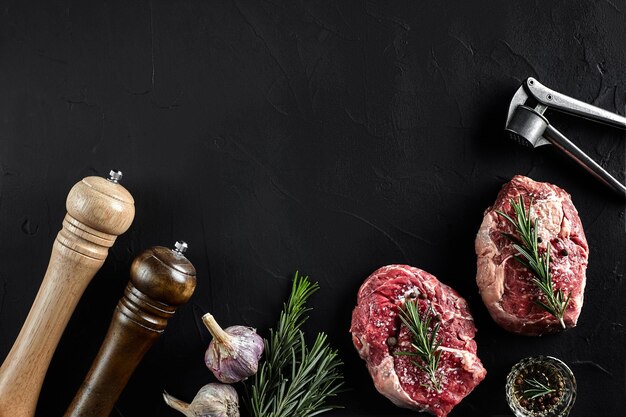 Deux steaks de faux-filet frais crus avec sel, poivre, romarin et ail sur fond noir