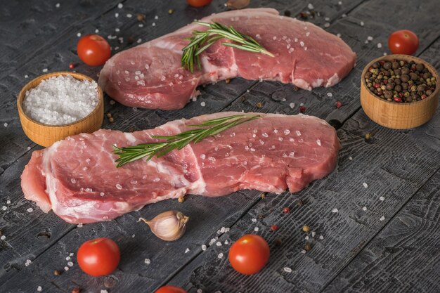 Deux steaks de porc aux assaisonnements et tomates cerises éparpillés sur une table en bois. Ingrédients pour la cuisson des plats de viande.