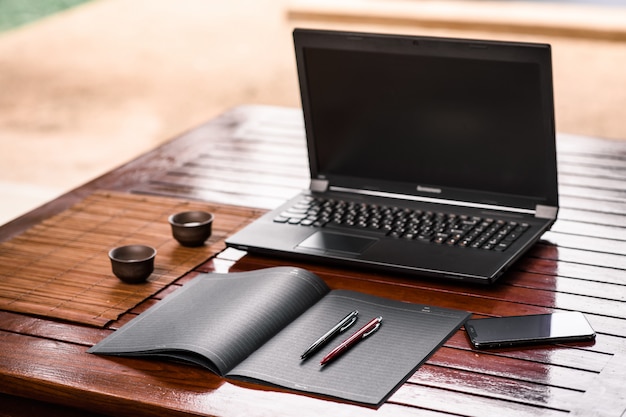 Deux stylos de bureau de couleur noir et rouge situés sur un cahier noir posé sur une table en bois sur laquelle il y a un ordinateur portable ouvert, un téléphone noir et deux tasses pour les boissons