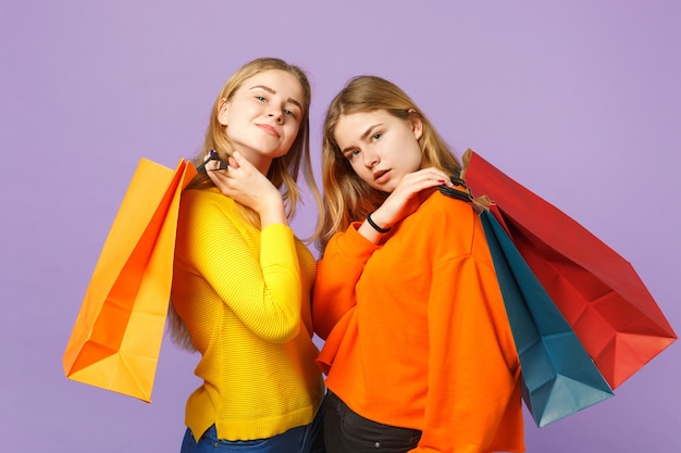 Deux superbes jeunes filles jumelles blondes vêtues de vêtements vifs tiennent un sac d'emballage avec des achats après le shopping isolés sur un mur bleu violet. Concept de mode de vie familial de personnes.